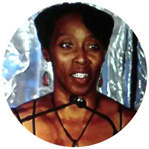 Gail Devers, wearing a patterned dress and red jewelry, is standing at a podium with multiple microphones, speaking in front of a metallic silver backdrop.