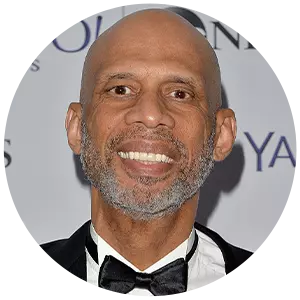 Kareem Abdul-Jabbar is dressed in a black tuxedo with a bow tie, smiling at an event with a backdrop featuring company logos.