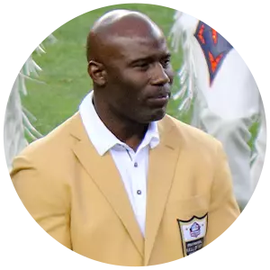 Terrell Davis is wearing a gold blazer with a Hall of Fame patch over a white collared shirt, standing on a sports field with hands clasped.