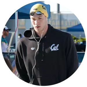 Ryan Murphy, wearing a yellow swim cap, goggles, and a black Cal hoodie, stands outside listening to earphones. There are people and tents in the background, and a young girl is in the foreground.