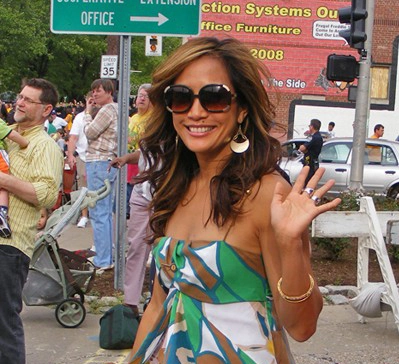 Carrie Ann Inaba seated and smiling, wearing a bright multicolored top and waving toward the camera.