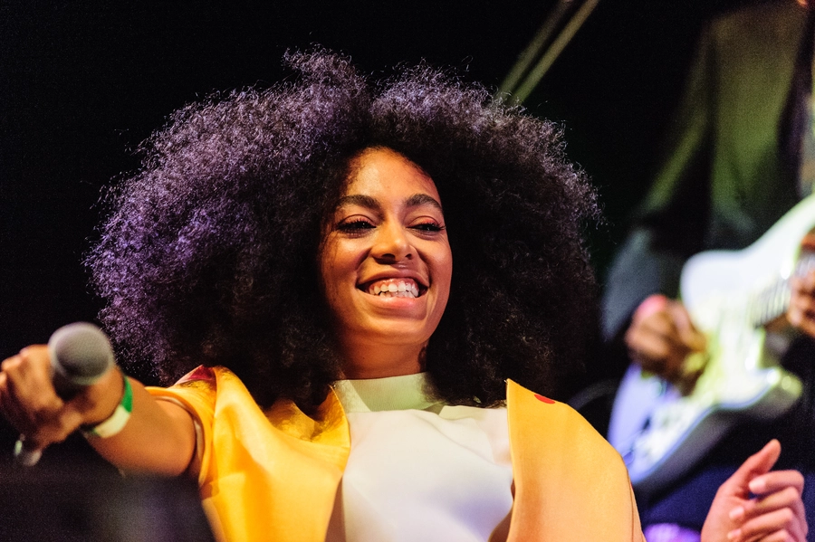 Solange Knowles smiling and holding a microphone, wearing a yellow and white outfit on stage.